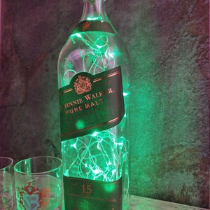 May include: A clear glass bottle with a green label that says "Johnnie Walker Pure Malt Scotch Whisky" and "15" is filled with green fairy lights. The bottle is sitting on a wooden surface next to two clear glass tumblers.