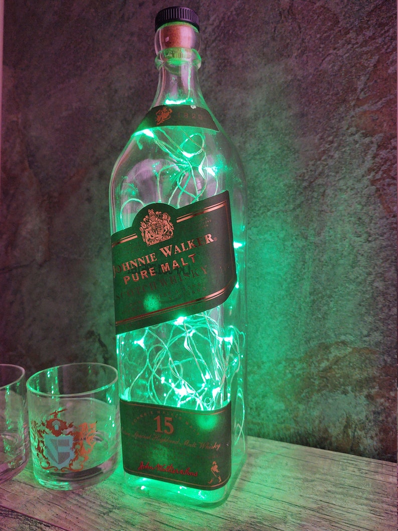 May include: A clear glass bottle with a green label that says "Johnnie Walker Pure Malt Scotch Whisky" and "15" is filled with green fairy lights. The bottle is sitting on a wooden surface next to two clear glass tumblers.