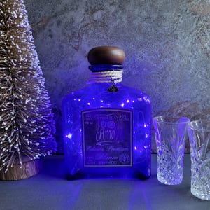 May include: A blue tequila bottle with a brown stopper, wrapped with string, and illuminated with blue lights. The bottle has a label that reads "El Amo Blanco" and is next to two clear shot glasses. A small decorative tree is on the left.