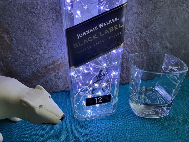 Puede incluir: Una botella de vidrio transparente con luces blancas de cuerda en su interior. La botella est&aacute; etiquetada con "Johnnie Walker Black Label Blended Scotch Whisky 12 Years Old". Un vaso de vidrio transparente se encuentra junto a la botella.