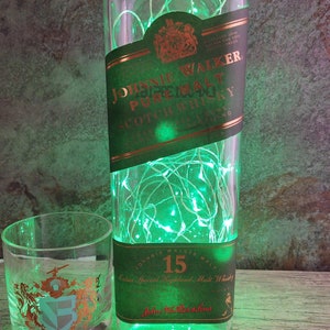 May include: A green glass bottle with a label that reads "Johnnie Walker Pure Malt Scotch Whisky" and a string of green lights inside. The bottle is sitting on a wooden surface next to two clear glass shot glasses.