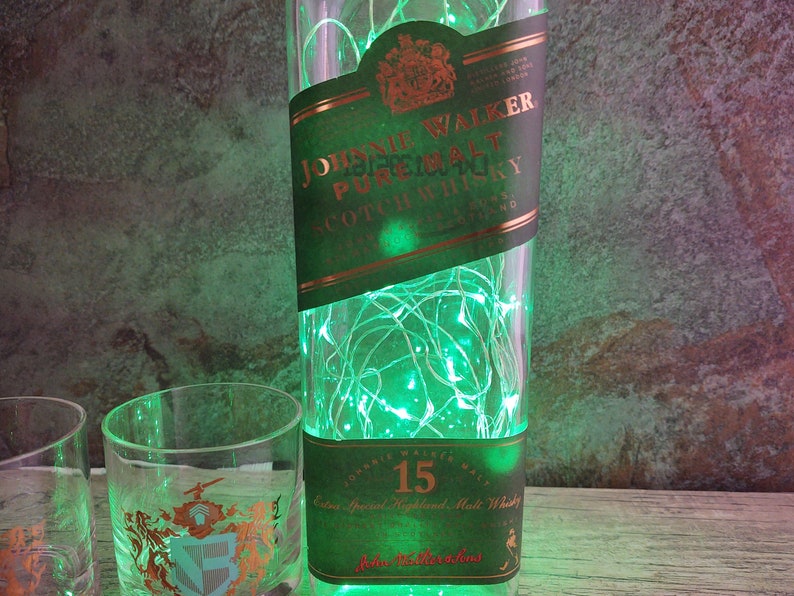 May include: A green glass bottle with a label that reads "Johnnie Walker Pure Malt Scotch Whisky" and a string of green lights inside. The bottle is sitting on a wooden surface next to two clear glass shot glasses.