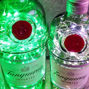 May include: Two clear glass bottles with a red label that says "Tanqueray Imported" and a red seal. The bottles are filled with white string lights. One bottle has green lights around it.