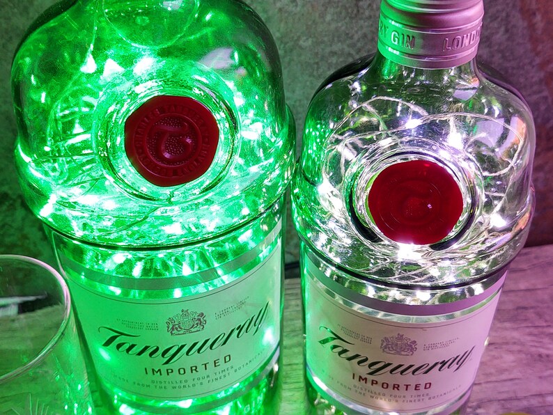 May include: Two clear glass bottles with a red label that says "Tanqueray Imported" and a red seal. The bottles are filled with white string lights. One bottle has green lights around it.