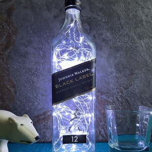 Puede incluir: Una botella de vidrio transparente con luces blancas de cuerda en su interior. La botella est&aacute; etiquetada con "Johnnie Walker Black Label Blended Scotch Whisky" y tiene una etiqueta negra con una silueta blanca de un hombre caminando. La botella tiene 12 a&ntilde;os de edad.