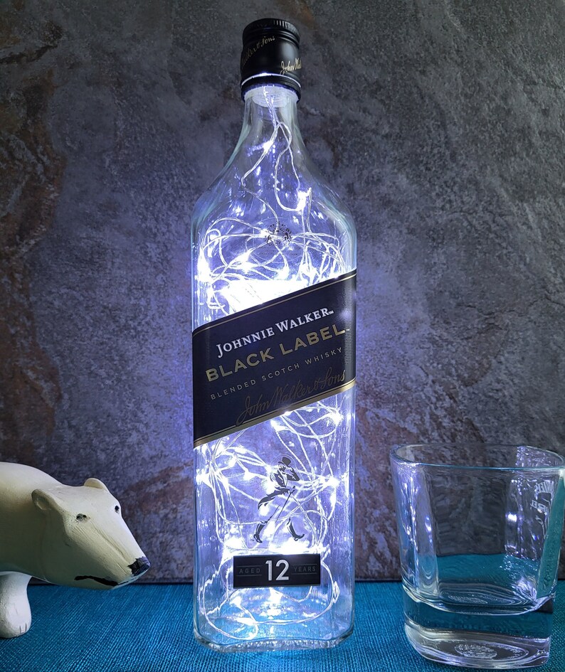 Puede incluir: Una botella de vidrio transparente con luces blancas de cuerda en su interior. La botella est&aacute; etiquetada con "Johnnie Walker Black Label Blended Scotch Whisky" y tiene una etiqueta negra con una silueta blanca de un hombre caminando. La botella tiene 12 a&ntilde;os de edad.