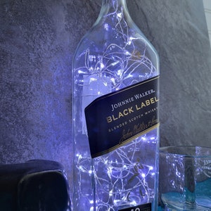 Puede incluir: Una botella de vidrio transparente con luces blancas en su interior. La botella tiene una etiqueta negra que dice "Johnnie Walker Black Label Blended Scotch Whisky". La botella est&aacute; sobre una superficie azul.