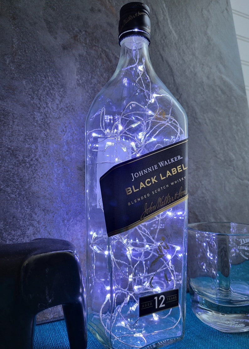 Puede incluir: Una botella de vidrio transparente con luces blancas en su interior. La botella tiene una etiqueta negra que dice "Johnnie Walker Black Label Blended Scotch Whisky". La botella est&aacute; sobre una superficie azul.