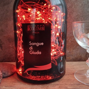 May include: A glass bottle with red string lights inside. The bottle has a black label with the text "Scarpa Colombi Italia" and "Sangue di Giuda".