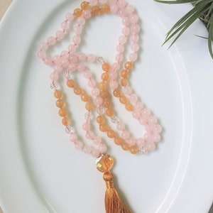 May include: A pink and yellow gemstone mala necklace with a brown tassel. The necklace is made with rose quartz and citrine beads.
