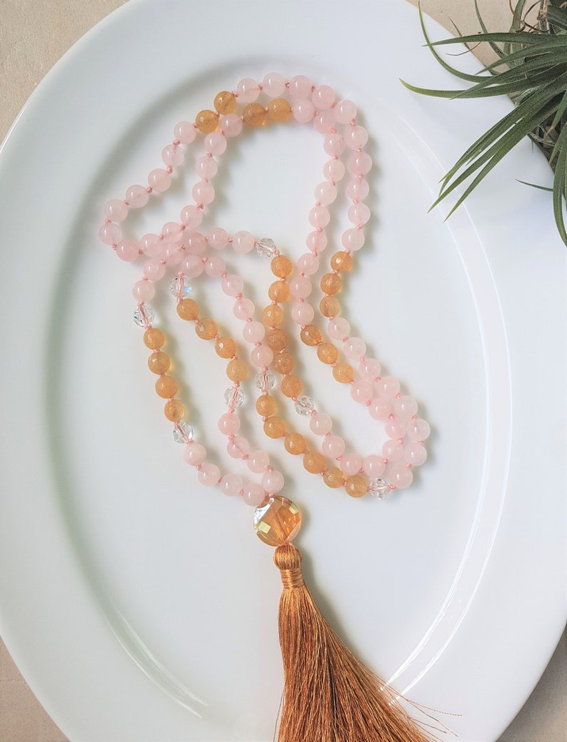 May include: A pink and yellow gemstone mala necklace with a brown tassel. The necklace is made with rose quartz and citrine beads.