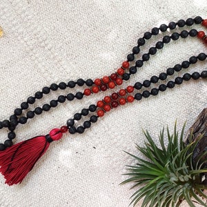 以下が含まれることがあります： 黒い溶岩石と赤いカーネリアンのビーズで作られたマラネックレスで、赤いタッセルが付いています。