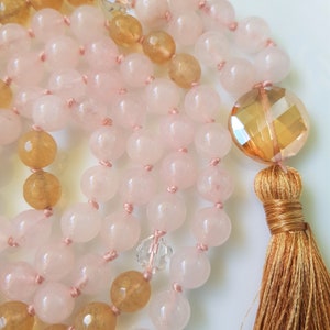 May include: A pink and yellow gemstone mala necklace with a tassel. The necklace is made with rose quartz and citrine beads.