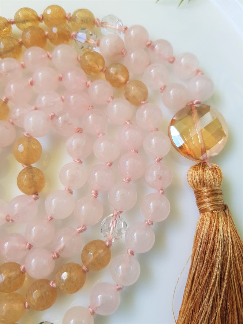 May include: A pink and yellow gemstone mala necklace with a tassel. The necklace is made with rose quartz and citrine beads.