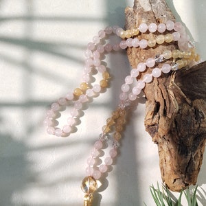 May include: A pink and yellow crystal mala necklace with a gold tassel. The necklace is draped over a piece of driftwood.