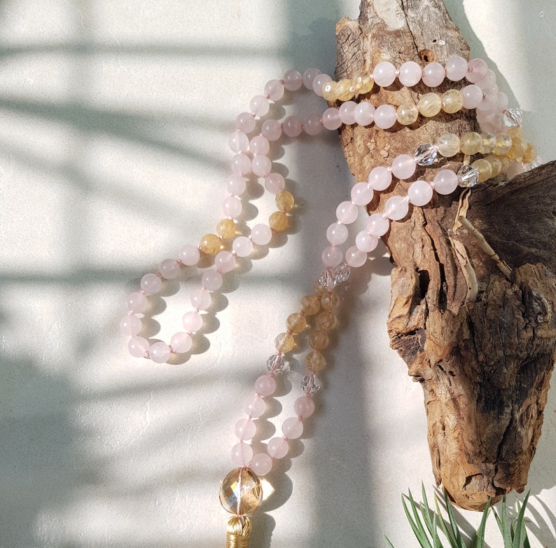 May include: A pink and yellow crystal mala necklace with a gold tassel. The necklace is draped over a piece of driftwood.