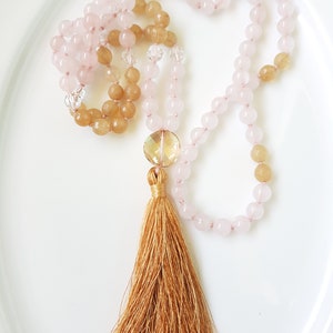 May include: A mala or prayer beads necklace with pink and brown beads and a gold tassel. The beads are strung on a clear cord.