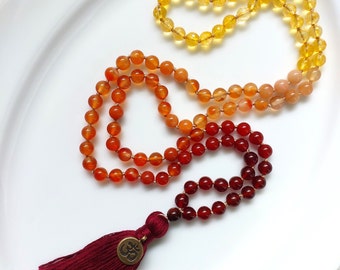 Puede incluir: Un collar mala de piedras preciosas multicolores con un borla roja. El collar está hecho de cuentas de cornalina, citrino y cuarzo transparente. La borla tiene un amuleto con el símbolo Om.