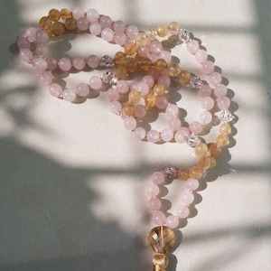 May include: A pink and yellow crystal mala necklace with a gold tassel. The necklace is made of rose quartz and citrine crystals.