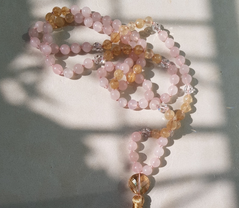 May include: A pink and yellow crystal mala necklace with a gold tassel. The necklace is made of rose quartz and citrine crystals.