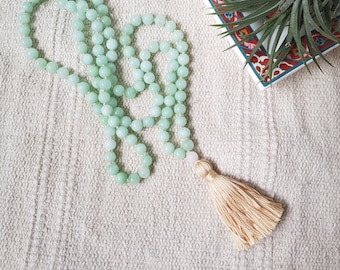 May include: A light green mala necklace with a tassel made of beige fabric. The necklace is made of round beads and is lying on a white textured surface.