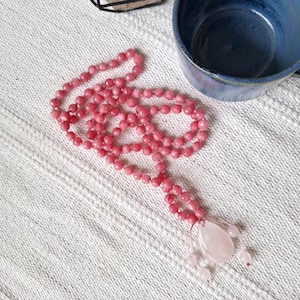 May include: A pink beaded necklace with a teardrop-shaped pendant and small round beads. The necklace is made of small, round, pink beads and a larger, polished pink stone pendant. The necklace is displayed on a white textured surface.