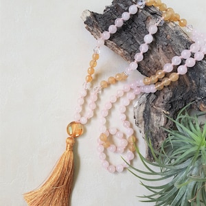May include: A pink and yellow gemstone mala with a tassel. The mala is made of round beads and is strung on a cord. The tassel is made of a brown fabric.