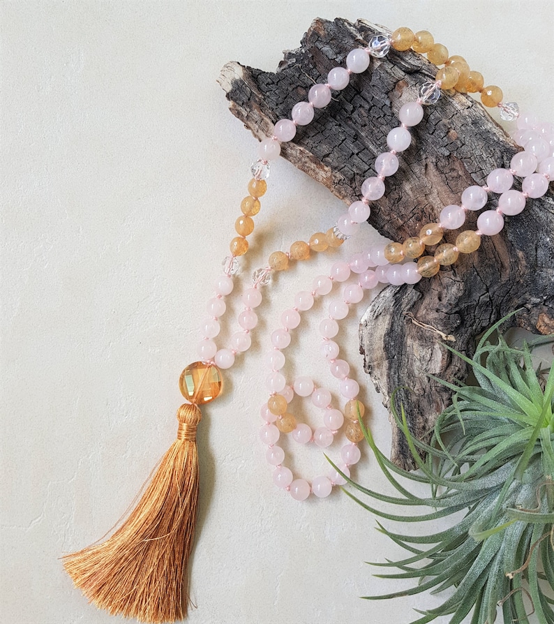 May include: A pink and yellow gemstone mala with a tassel. The mala is made of round beads and is strung on a cord. The tassel is made of a brown fabric.