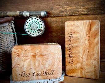 Puede incluir: Caja de aparejos de pesca con mosca de madera y tabla de cortar de madera con el texto "The Catskill" grabado. Un carrete y una caña de pescar son visibles en el fondo. La caja de aparejos está sobre un tapete blanco.