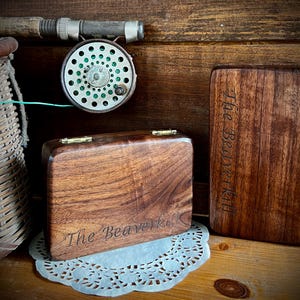 May include: A wooden fly fishing reel and two wooden boxes with the text "The Beaverkill" engraved on them. The boxes have brass hinges and clasps. A wicker fishing creel and a fishing rod are also visible.