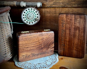 Puede incluir: Un carrete de pesca con mosca y dos cajas de madera con el texto "The Beaverkill" grabado. Las cajas tienen bisagras y cierres de latón. También se ve un esportón de pesca de mimbre y una caña de pescar.