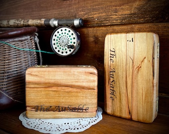 Puede incluir: Dos cajas de madera con el texto "The AuSable" grabado. Una caja es rectangular y la otra cuadrada. Un carrete y una caña de pescar son visibles en el fondo. Las cajas son de madera clara con veta natural.