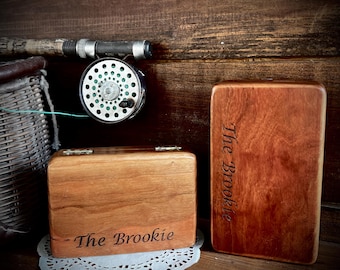 Puede incluir: Una caja de madera y una placa rectangular de madera con el texto "The Brookie" grabado. Un carrete y una caña de pescar son visibles en el fondo. La caja está sobre un mantelito blanco.