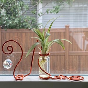 May include: A copper-colored wire plant stand with a glass bottle holding a green plant. A clear crystal prism hangs from the stand. The stand is on a white windowsill with a blurred background of a wooden fence and trees.