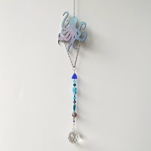 May include: A hanging octopus-shaped suncatcher with a gradient of blue and purple hues. The octopus is connected to a chain with blue and green beads, and a clear crystal ball at the bottom. The suncatcher is designed to hang from a hook.
