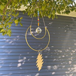 May include: A decorative suncatcher with a gold-colored metal frame in a crescent moon shape, a faceted crystal ball, and a gold lightning bolt charm. The suncatcher hangs from a chain with colorful beads. The background is a blue wall and green foliage.