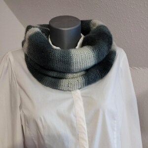 May include: A knitted cowl scarf in grey and blue tones, displayed on a mannequin. The scarf features a striped pattern and is worn over a white collared shirt. The cowl is a seamless loop, designed to be worn around the neck.