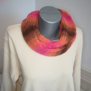 Op de afbeelding: Een gebreide oneindige sjaal in tinten bruin, oranje en roze. De sjaal wordt getoond op een wit mannequin.