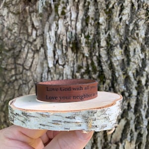 Puede incluir: Pulsera de cuero marrón con el texto "Ama a Dios con todo tu corazón. Ama a tu prójimo como a ti mismo." grabado en ella.