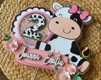 May include: A cow-themed birthday cake topper with the number 2, a pink bow, and the name "Joanna Emilia." The topper features a black and white cow design, pink flowers, and a clear acrylic stick for insertion into a cake.