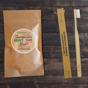 May include: A brown paper bag with a label that reads "120 Dentin Toothpaste Mint Tabs with Fluoride 100% Biodegradable Packaging Planet Wood". Next to the bag is a brown cardboard box with the text "The Eco-Friendly Bamboo Toothbrush Planet Wood". A bamboo toothbrush is next to the box.