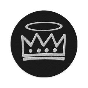Puede incluir: Un parche circular negro con una corona y un halo bordados en blanco. La corona tiene tres puntas y tres círculos en el centro.