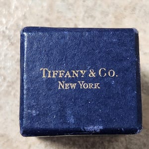 May include: A small, square, navy blue box with the words "TIFFANY & CO. NEW YORK" in gold lettering. The box has a textured surface and appears to be made of a soft material. The box is slightly worn.