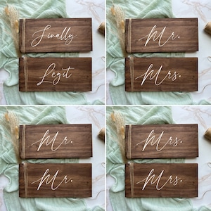 May include: A selection of dark brown wooden signs with white calligraphy. Some signs read "Finally" and "Legit", while others say "Mr." and "Mrs." in elegant script. Each sign is wrapped with twine.