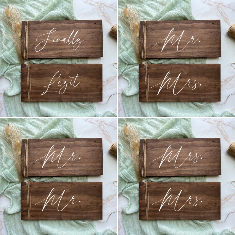 May include: A selection of dark brown wooden signs with white calligraphy. Some signs read "Finally" and "Legit", while others say "Mr." and "Mrs." in elegant script. Each sign is wrapped with twine.