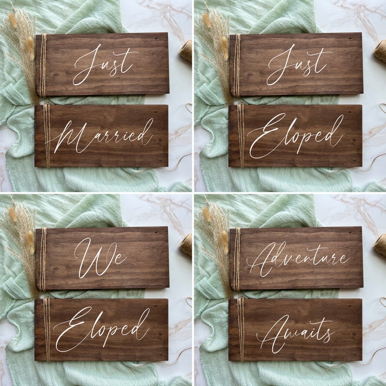 May include: Six dark brown wooden signs with white cursive text, decorated with twine. The signs display phrases such as "Just," "Married," "Eloped," "We," "Adventure," and "Awaits." They are arranged on a light green fabric background.