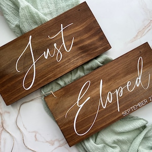 May include: Two rectangular wooden signs with white cursive text. One sign reads "Just", the other reads "Eloped September 21". The signs are stained a dark brown and have a rustic aesthetic, ideal for a wedding.