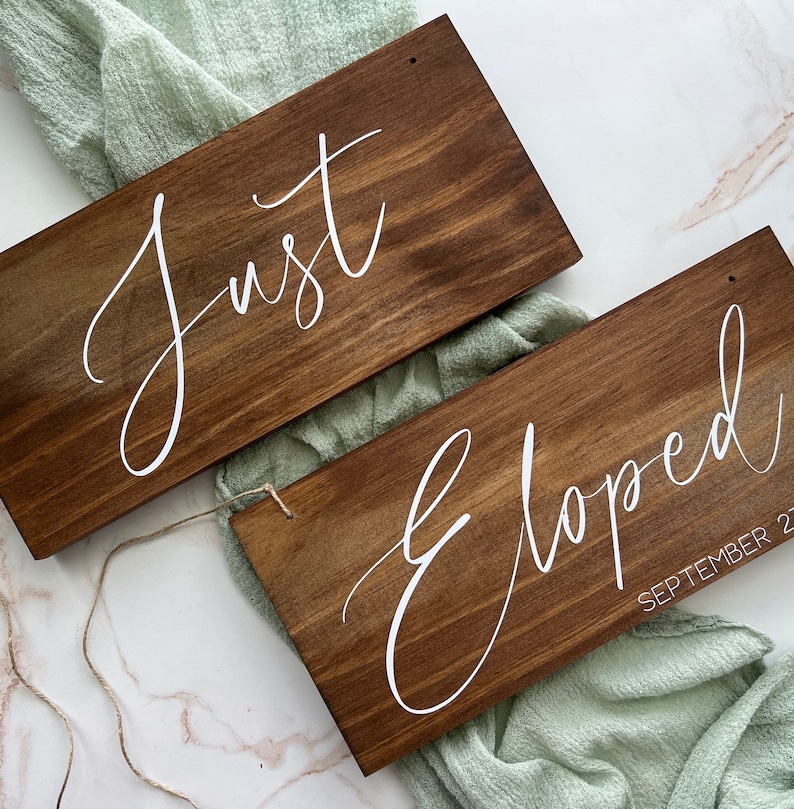 May include: Two rectangular wooden signs with white cursive text. One sign reads "Just", the other reads "Eloped September 21". The signs are stained a dark brown and have a rustic aesthetic, ideal for a wedding.