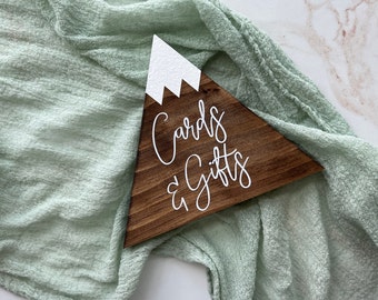 May include: A wooden sign shaped like a mountain with a white snow-capped peak. The sign says "Cards & Gifts" in white cursive lettering.