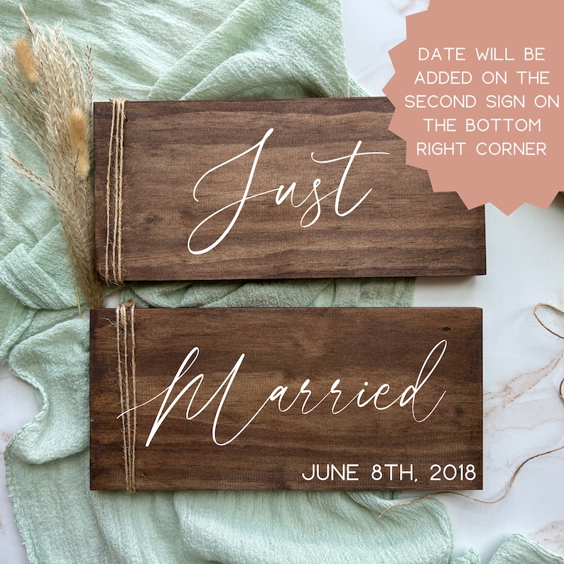 May include: Two rectangular wooden signs with the words "Just" and "Married" in white script. The "Married" sign includes "June 8th, 2018". Each sign is wrapped with twine. The background is a light green fabric.